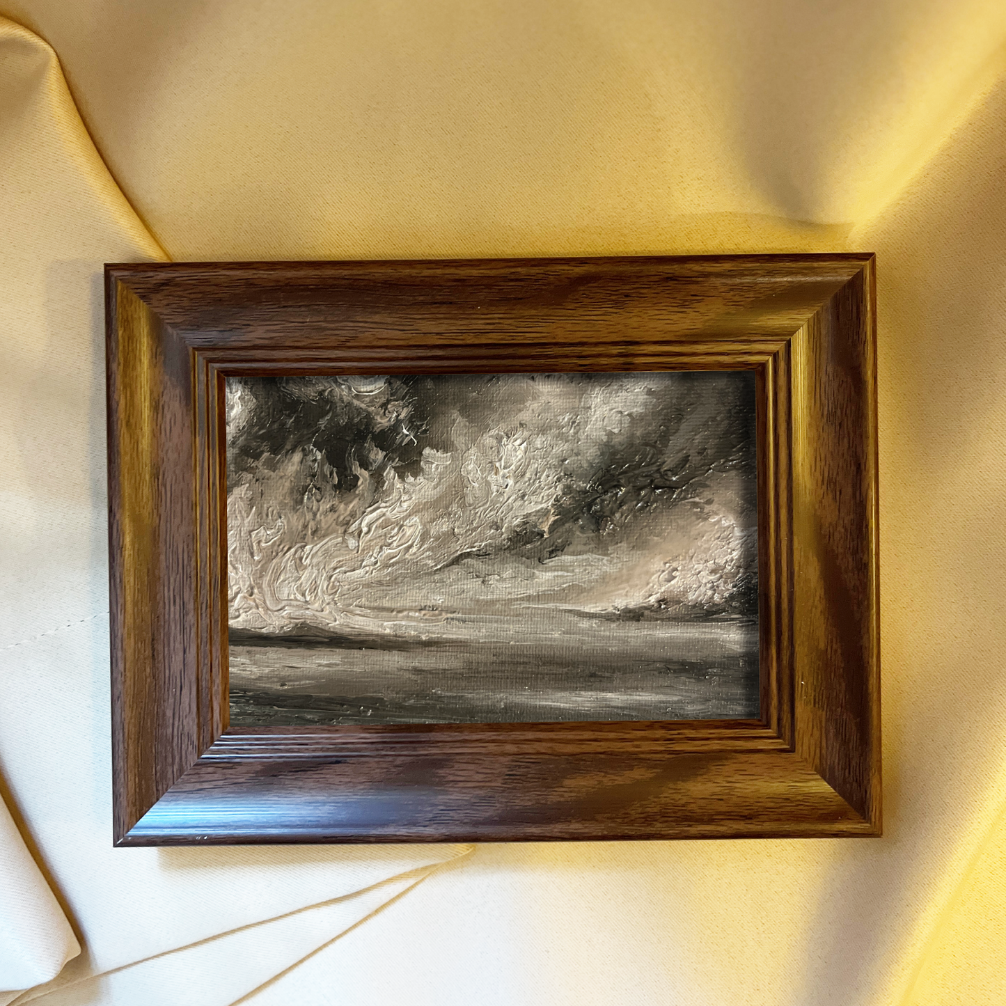 A framed landscape painting depicting a dramatic stormy sky over a body of water, housed in a dark brown wooden frame and displayed on a light beige fabric background.