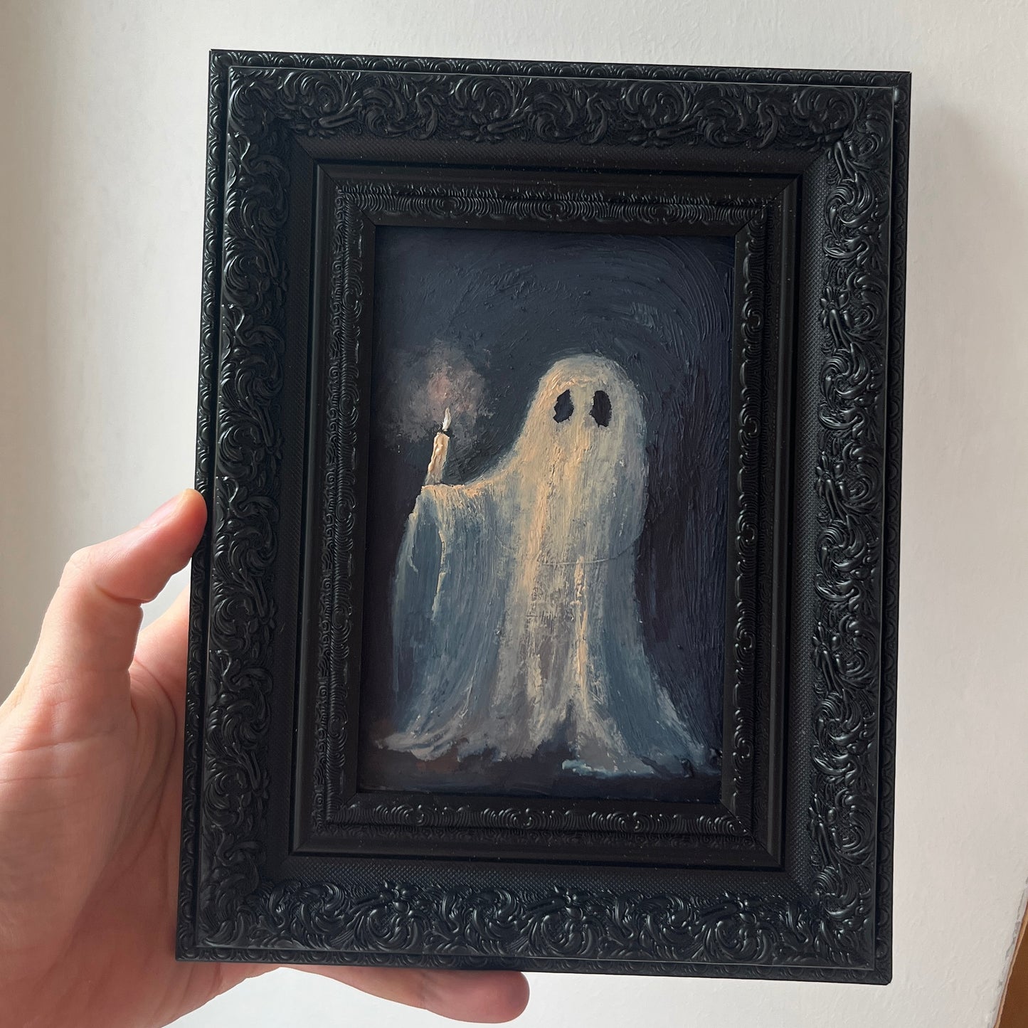 Framed artwork of a ghost holding a candle, held by a hand against a neutral background