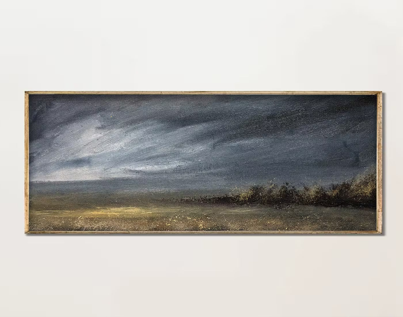 A framed landscape painting featuring a dramatic dark sky over a grassy field with a line of trees, displayed on a light neutral wall.